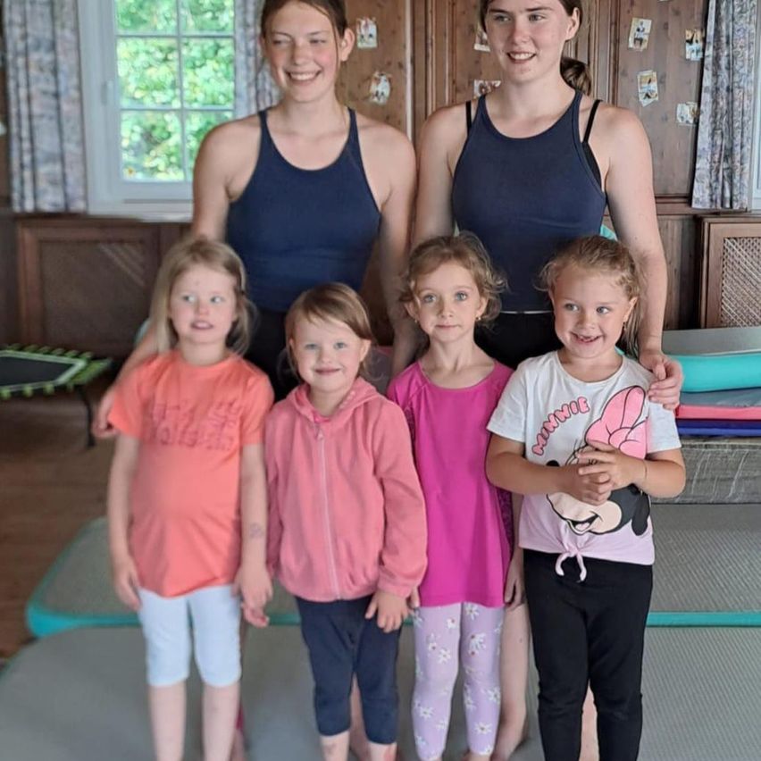 Zwei Frauen in Tanktops stehen mit vier jungen Mädchen und lächeln für ein Foto. Sie sind in einem Raum mit Holzwänden und einem Fenster mit Vorhängen. Ein Trampolin steht auf dem Boden hinter ihnen.