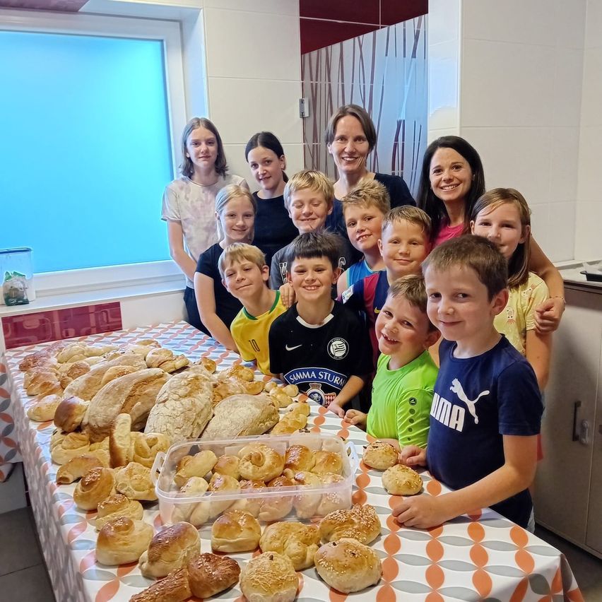 Eine Gruppe von Kindern und Erwachsenen versammelt sich um einen Tisch mit Brot und Gebäck und lächelt für ein Foto in einem Raum mit einem blauen Fenster und gefliesten Wänden.