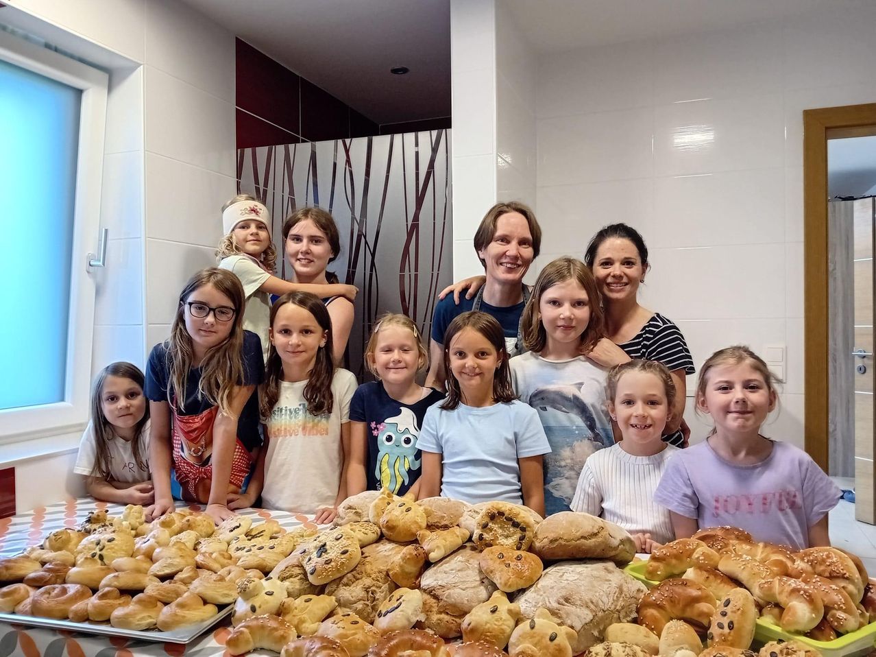 Eine Gruppe junger Mädchen und Erwachsener posiert für ein Foto in einem Raum. Sie lächeln und stehen um einen Tisch herum, der mit verschiedenen Broten gefüllt ist.