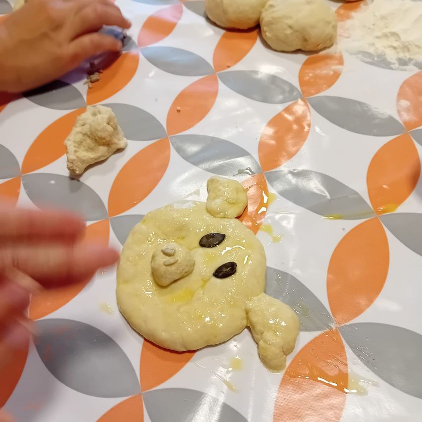 Eine Person formt einen Bären aus Teig. Der Teig liegt auf einem Tisch, der mit einem gemusterten Tuch bedeckt ist. Der Teig ist gelb und hat einige Samen darauf. Etwas Teig ist auf dem Tisch verstreut.