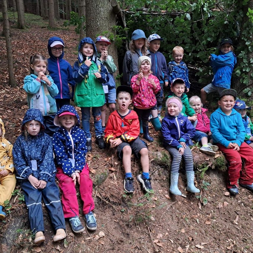Eine Gruppe von Kindern in Regenjacken sitzt auf einem Waldhang, einige mit offenem Mund, andere halten etwas zu essen.