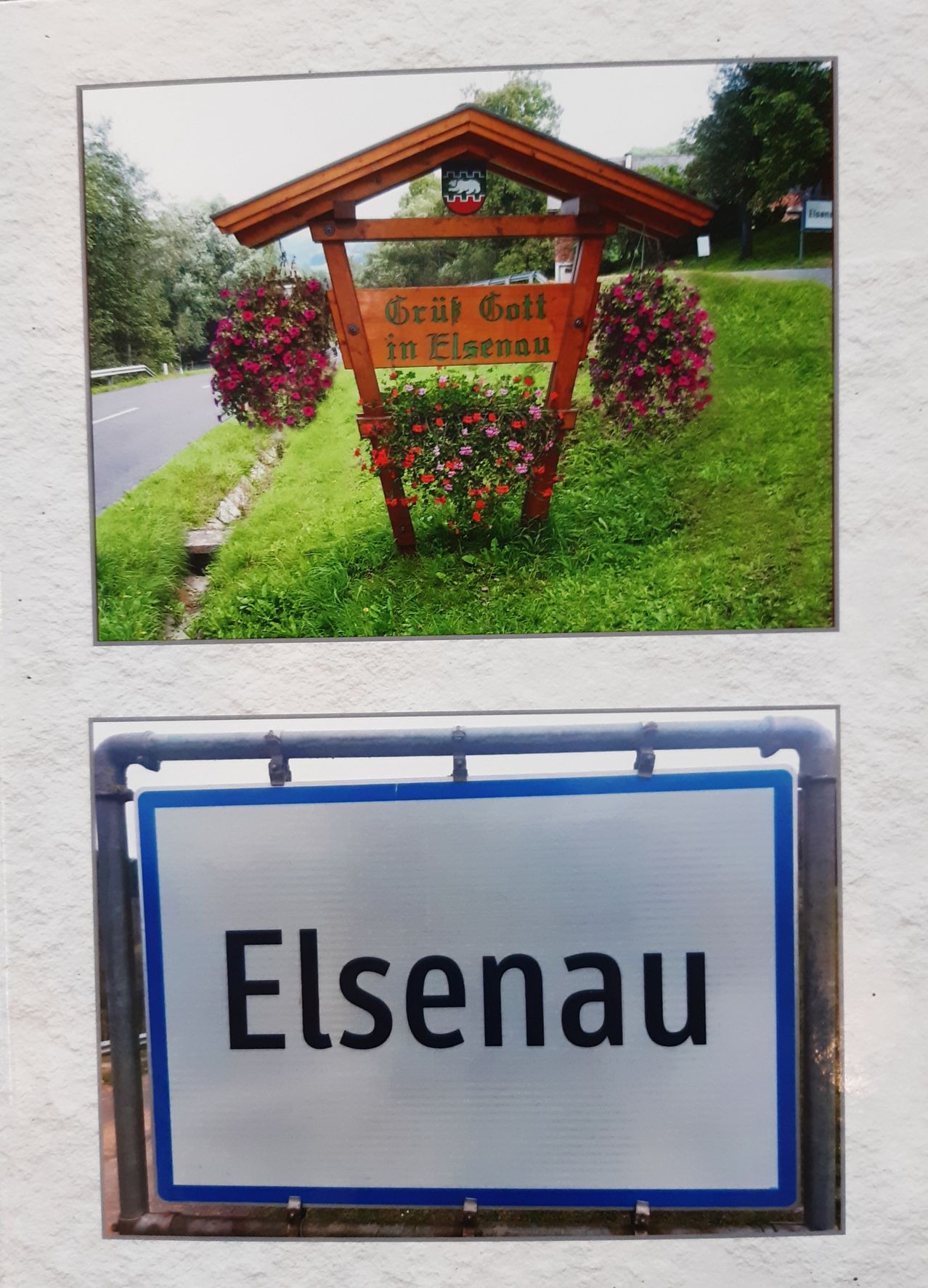 Ein hölzernes Schild mit Blumen und den Worten 'Gruf Gott in Elsennau' ist auf einem Rasen montiert. Eine Straße mit einem Geländer und Bäumen befindet sich daneben. Ein blaues Schild mit dem Wort 'Elsenau' ist unter dem Schild angebracht.
