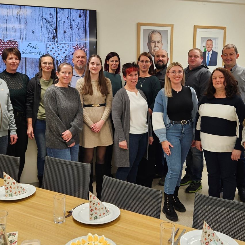 Eine Gruppe von Menschen steht in einem Raum. Ein Tisch hat Teller, Gläser und Besteck. Dahinter hängen zwei Bilderrahmen an der Wand. Der Text 'Frohe Weihnachten' ist auf dem Bildschirm.
