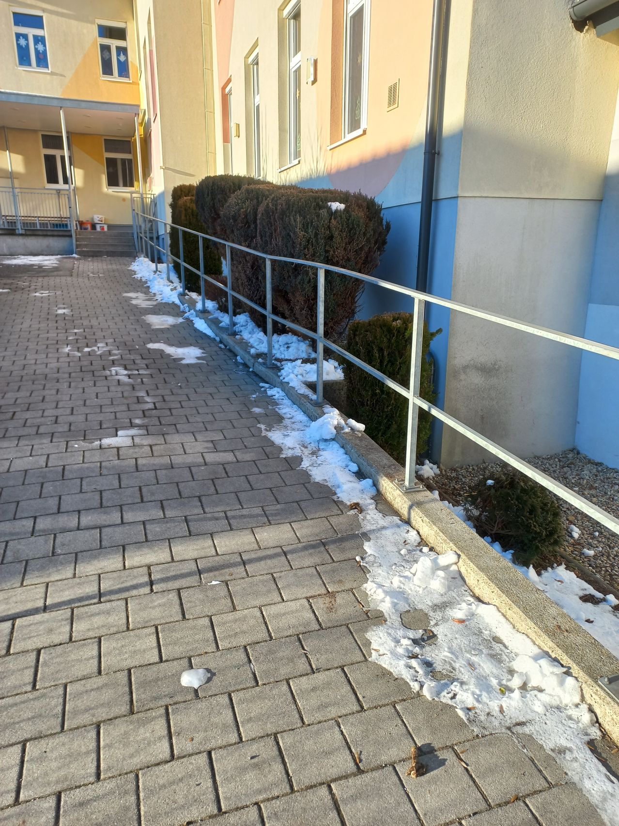 Ein gepflasterter Weg mit Schneeflecken neben einem Gebäude, mit einem Metallgeländer und einer Hecke.