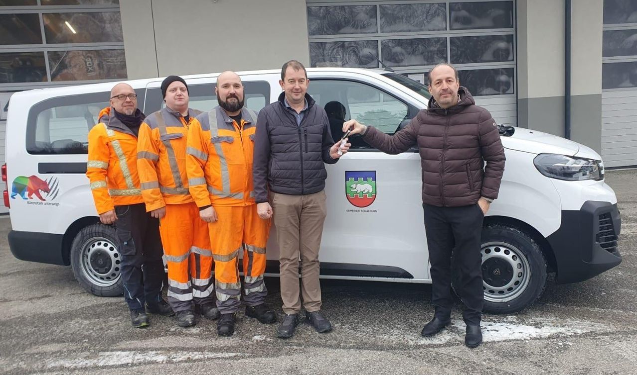 Five men are standing in front of a white van with a logo. One man holds a key, while the others are dressed in orange uniforms. They are likely employees of a utility company.