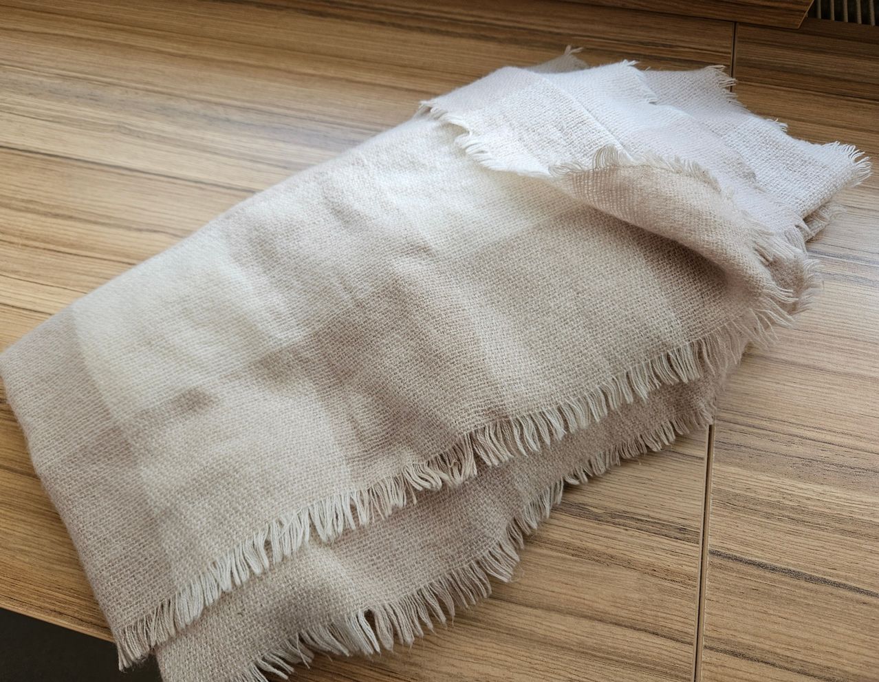 Ein beige-weiß karierter Schal mit ausgefransten Kanten liegt auf einer Holzoberfläche. Der Schal hat einen leicht rustikalen Look mit sichtbarer Textur.