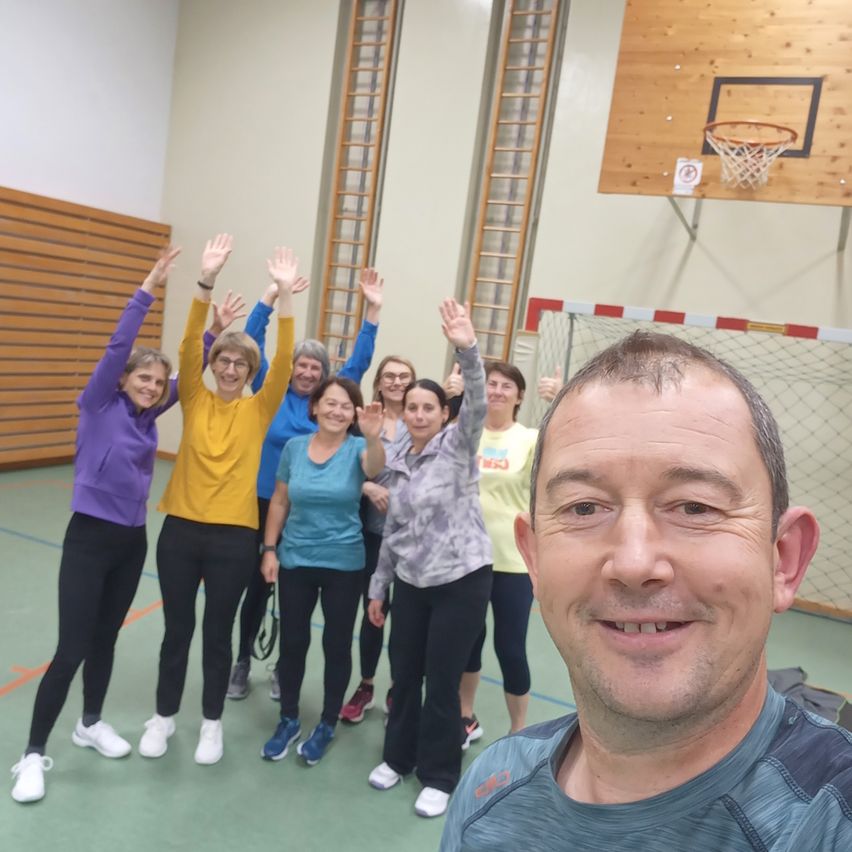 Eine Gruppe von Frauen in Trainingskleidung posiert für ein Foto in einer Turnhalle mit einem Basketballkorb im Hintergrund. Ein Mann lächelt davor.