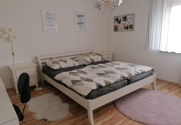A bedroom with a bed, wooden floor, and white walls. The bed has a quilt and pillows. Two nightstands and a chair are beside the bed. A lamp is on the ceiling.