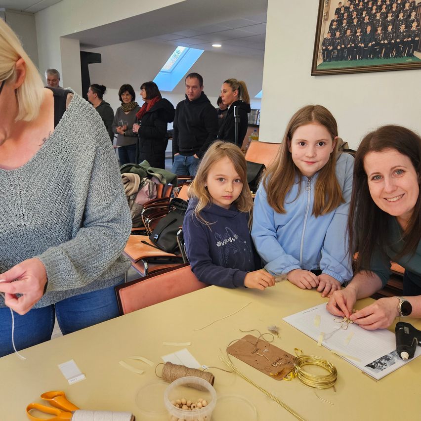 A woman with a tattoo and two girls are crafting at a table, with another woman beside them. Behind them, a group of people stand, and a framed picture is on the wall.