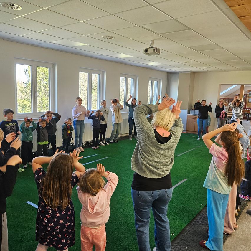 In a room with artificial turf, children and adults participate in a coordinated exercise session. The instructor leads with raised hands, while participants mimic her actions. The room features large windows with a view of trees outside.