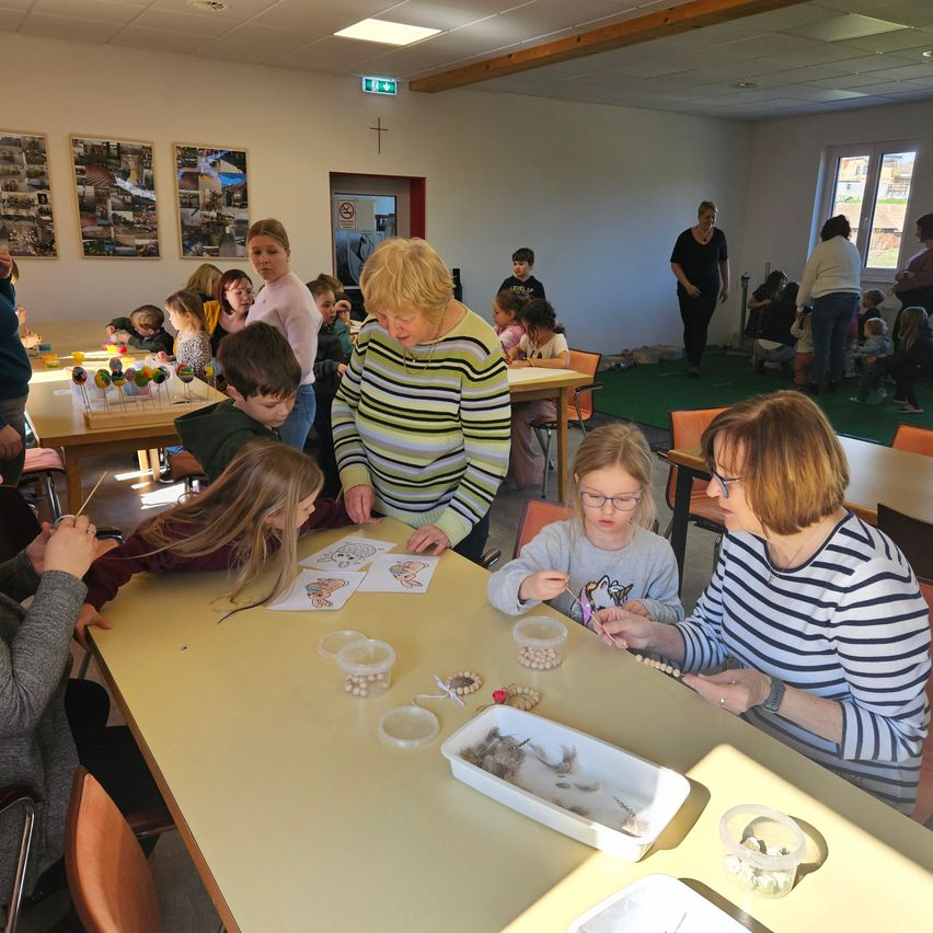 A group of children and adults are doing an activity at a table. They have papers and containers of beads. Some adults are helping them.