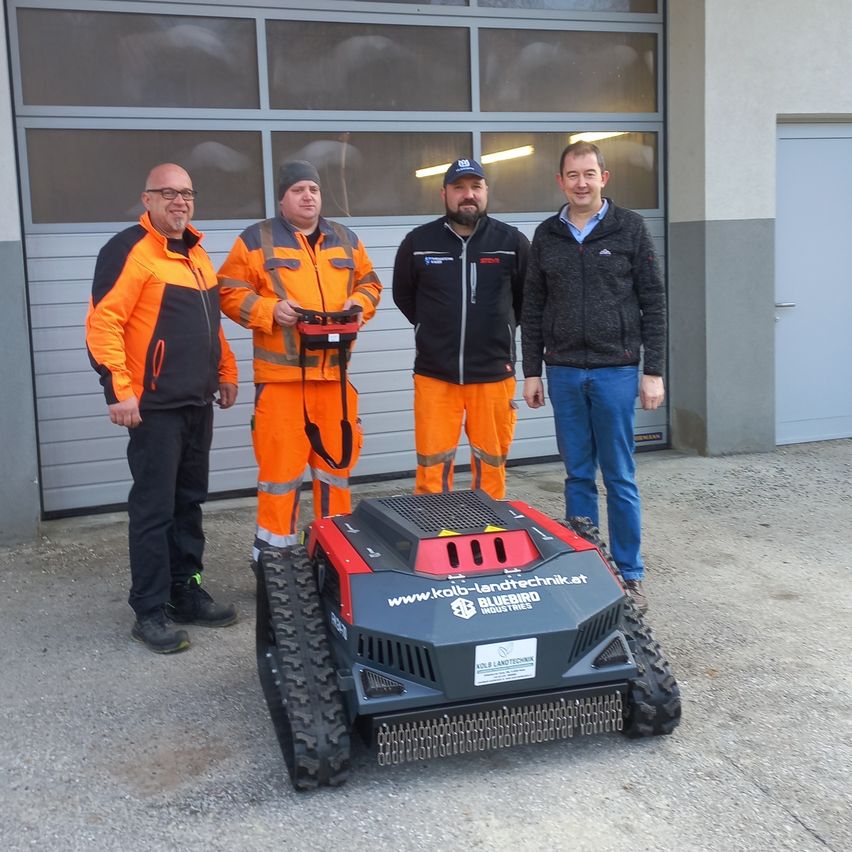 Vier Männer stehen vor einem Gebäude, zwei von ihnen tragen Schutzanzüge und die anderen beiden tragen Jacken. Sie stehen neben einem rot-schwarzen Roboter.