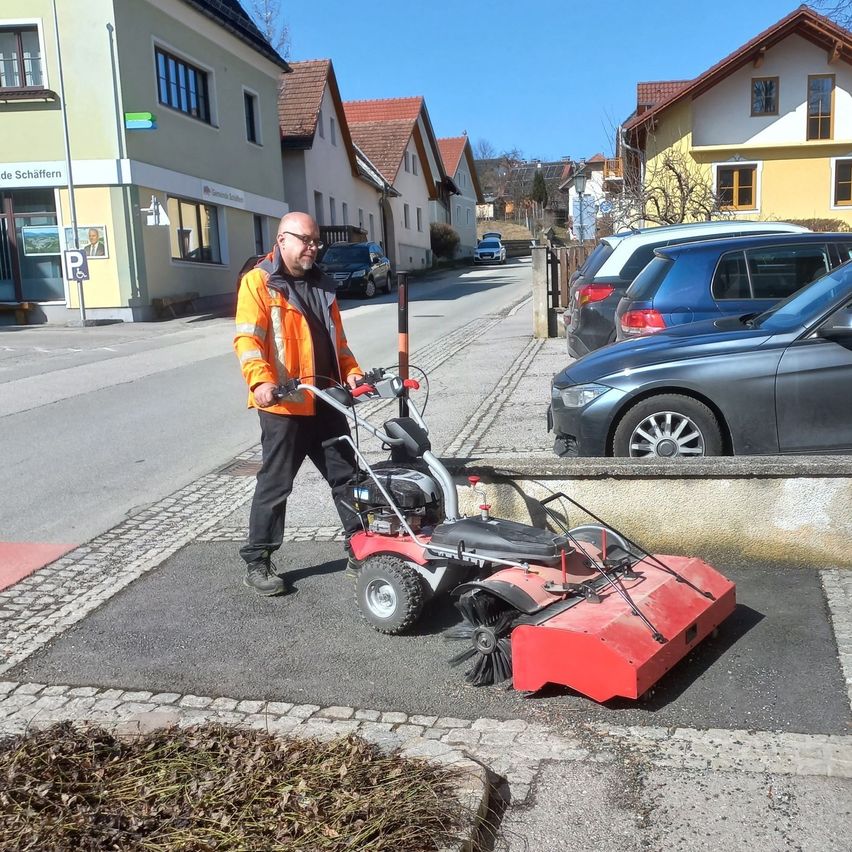 Ein Mann in einer orangefarbenen Jacke benutzt eine rote Straßenkehrmaschine auf einem Bürgersteig. Es gibt Häuser und geparkte Autos am Straßenrand.