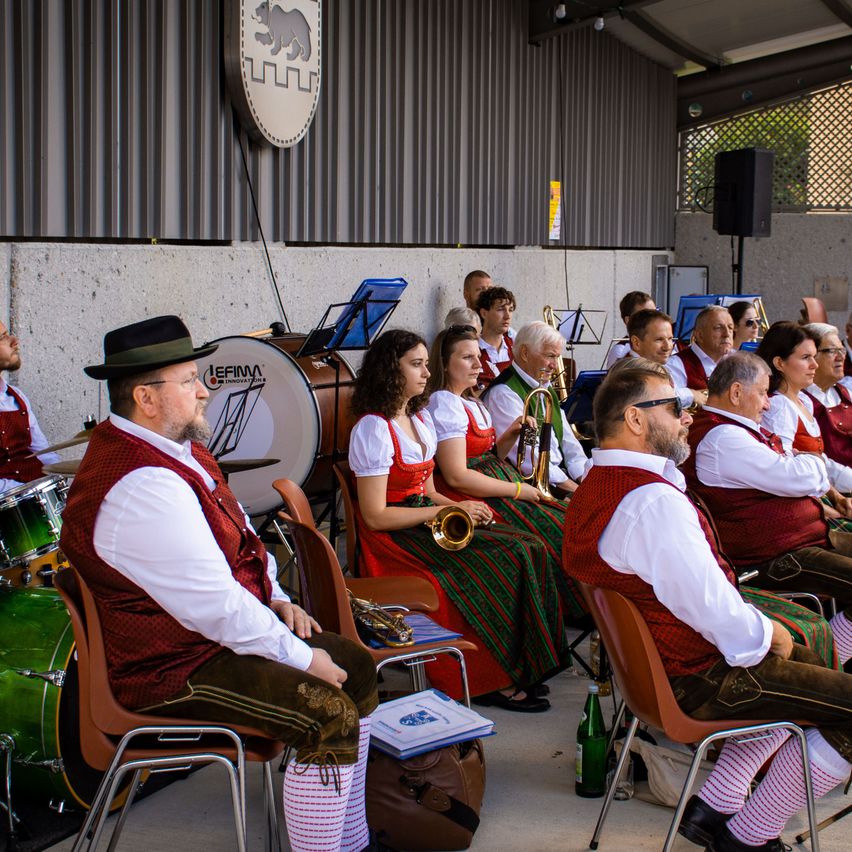 Eine Gruppe von Musikern in traditioneller Kleidung sitzt auf Stühlen, einige halten Instrumente. Ein Schlagzeug und ein Lautsprecher sind im Hintergrund zu sehen.