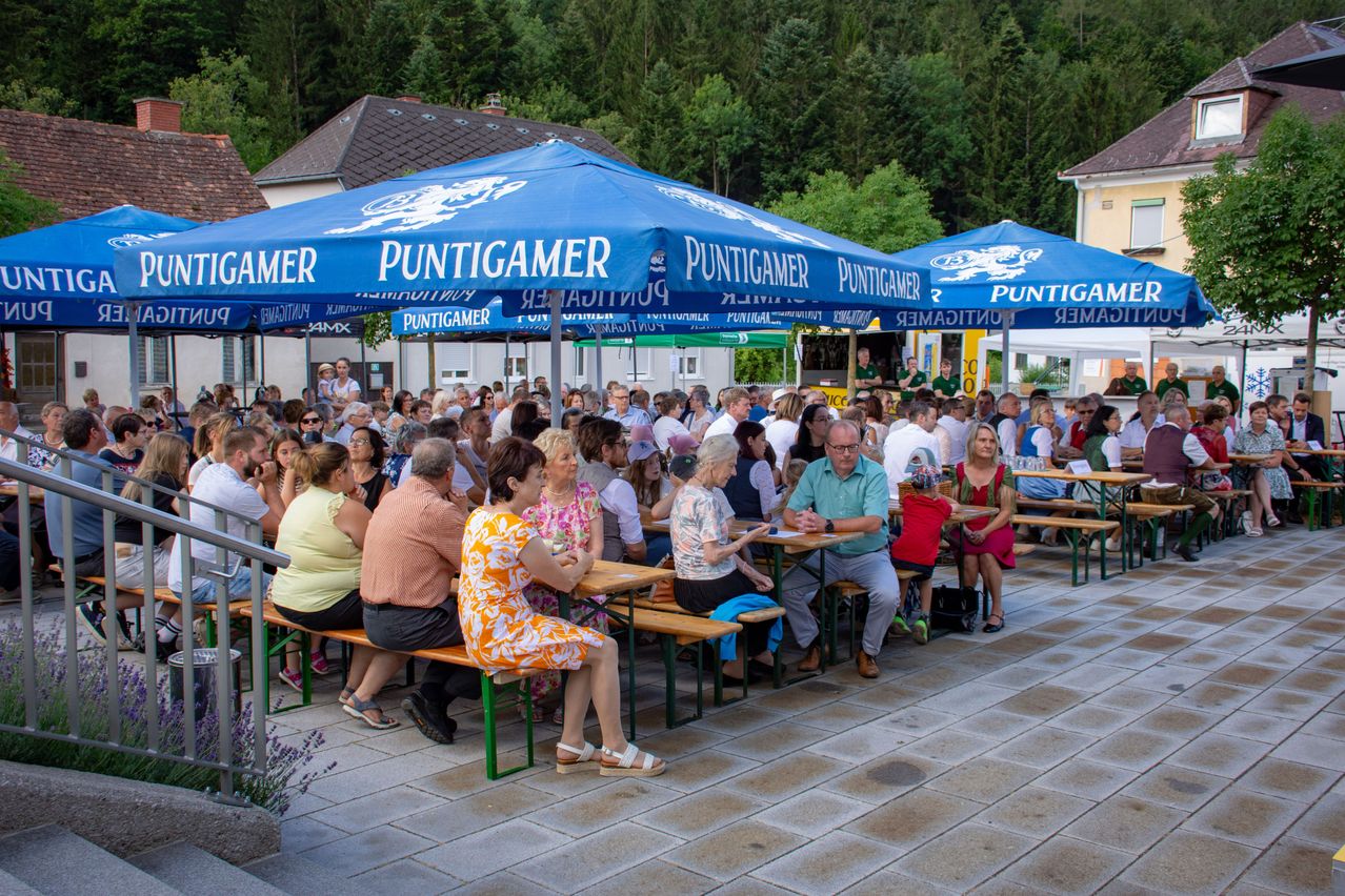 Eine große Menschenmenge versammelt sich unter blauen Zelten bei einer Outdoor-Veranstaltung. Viele sitzen an Tischen, während andere stehen und zusehen. Die Zelte tragen den Schriftzug 'Puntigamer'. Im Hintergrund befinden sich Bäume und ein Gebäude.