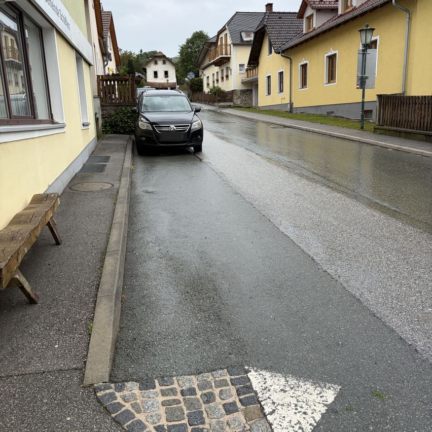 Ein schwarzes Auto ist auf einer nassen Straße geparkt, in der Nähe einer Holzbank und eines gelben Gebäudes. Die Straße ist von Häusern und einer Straßenlaterne gesäumt. Der Himmel ist bedeckt.