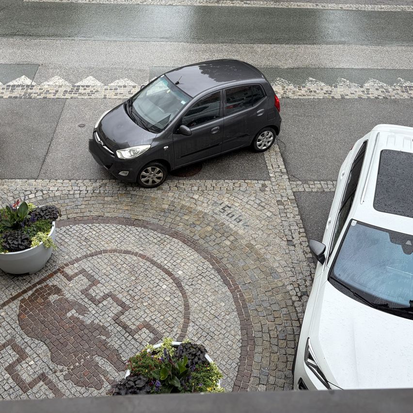 Eine oberirdische Ansicht eines Parkplatzes mit zwei Autos, eines schwarz und eines weiß. Das schwarze Auto steht in der Nähe eines steingepflasterten Bereichs mit Pflanzen. Das weiße Auto steht rechts.