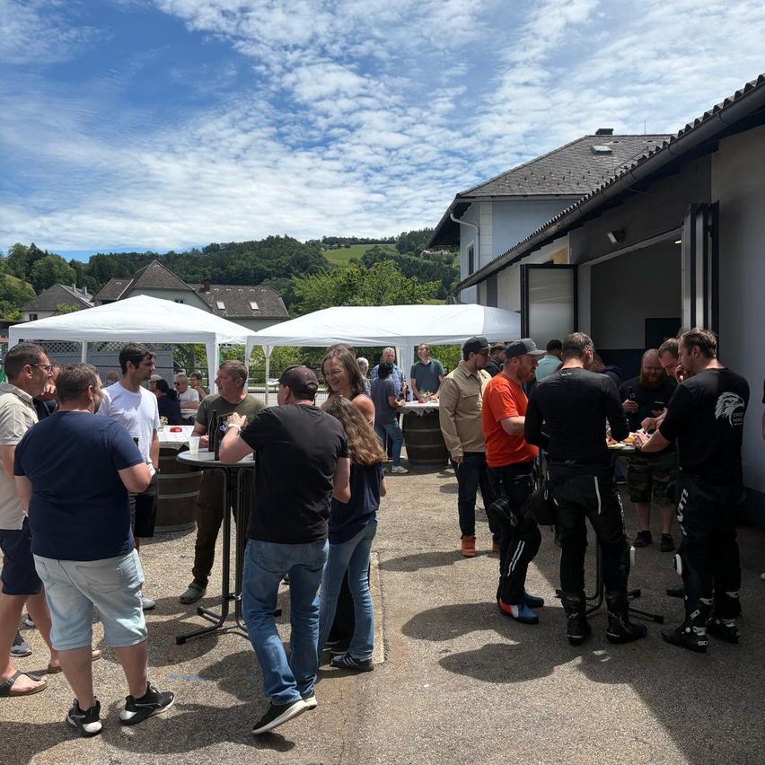 Eine Gruppe von Menschen steht unter Zelten in der Nähe eines Gebäudes, genießt Essen und Unterhaltung. Im Hintergrund befinden sich ein Haus und Berge unter einem blauen Himmel mit Wolken.