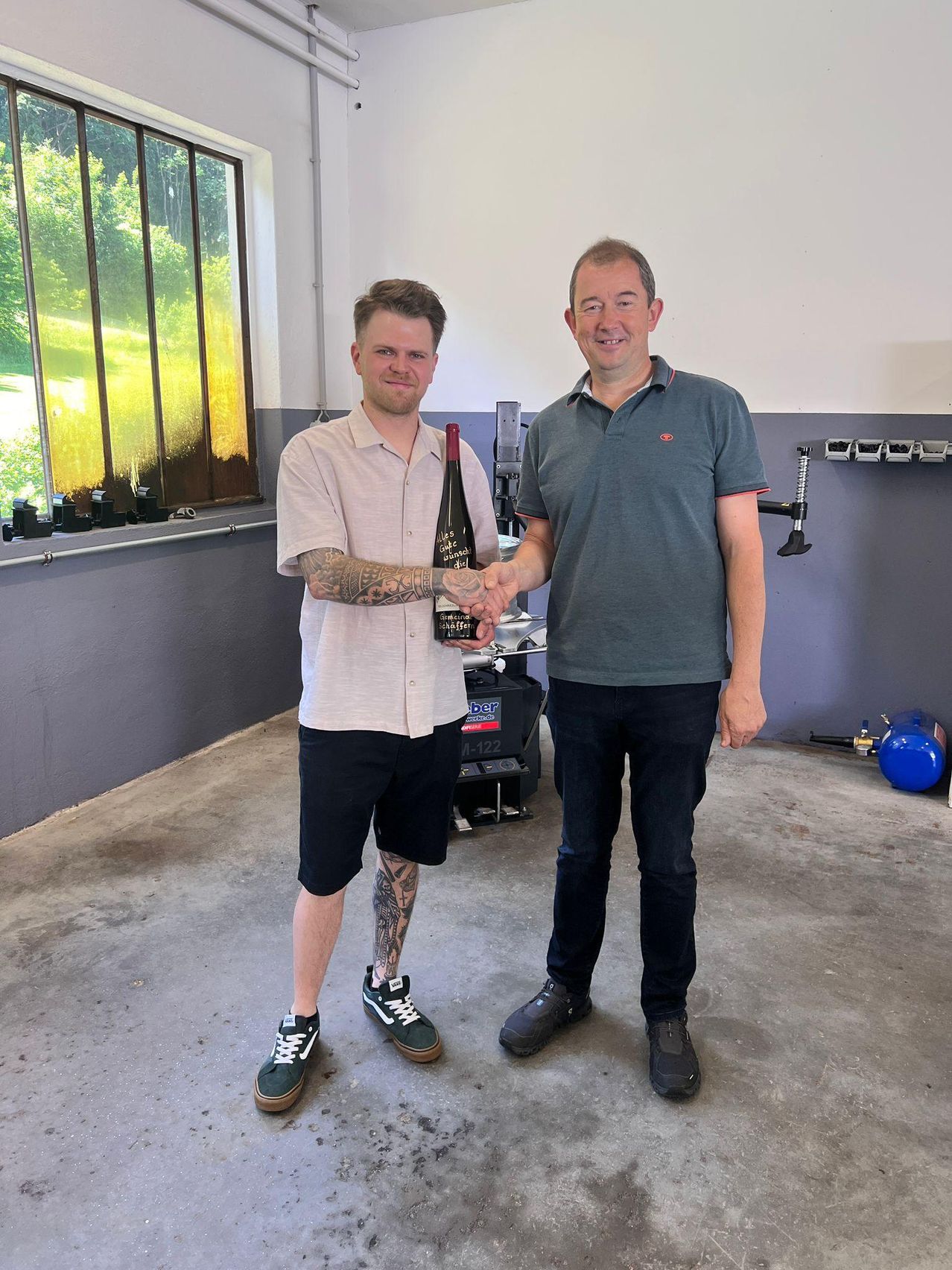 Zwei Männer in einer Garage, einer hält eine Flasche, beide lächeln. Ein Mann hat Tattoos und trägt Turnschuhe. Der andere trägt ein grünes Poloshirt und dunkle Hosen.