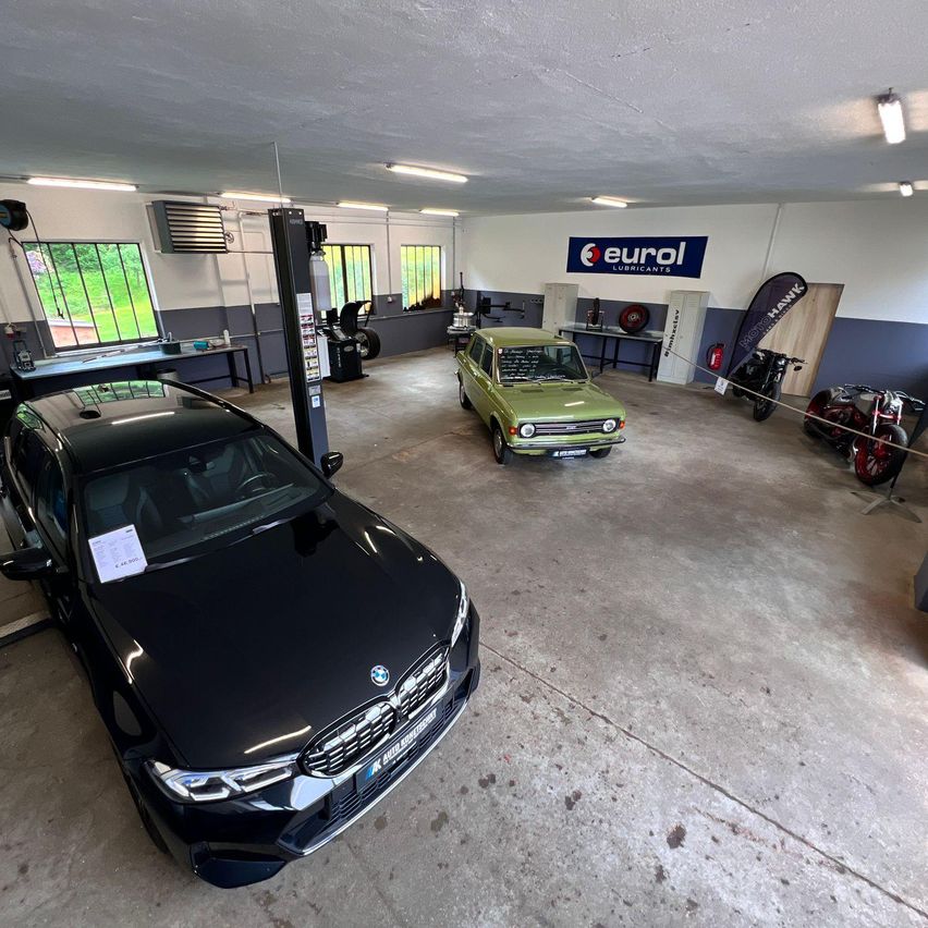 Eine Garage mit mehreren Fahrzeugen, einschließlich eines BMW, drinnen geparkt. Es sind Banner und Ausrüstung sichtbar.