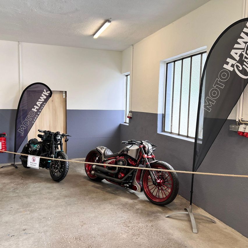 Zwei Motorräder sind in einer Garage ausgestellt. Eines ist ein schwarzer Chopper, das andere ist ein rotes Motorrad. Beide sind von Bannern umgeben.