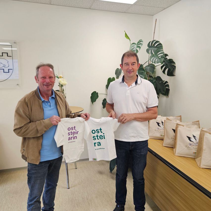Zwei Männer halten weiße T-Shirts mit den Worten 'Oststeierara' in grünem Text. Sie stehen in einem Büroraum mit einer Pflanze und Taschen auf einem Schreibtisch.
