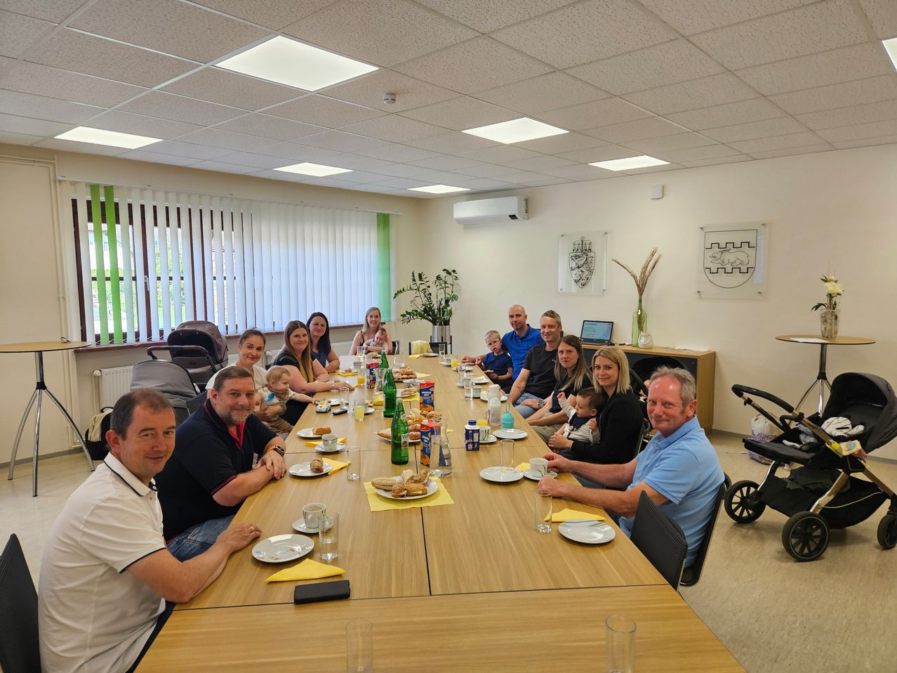 Eine Gruppe von Menschen sitzt um einen langen Tisch in einem Raum versammelt, wahrscheinlich für ein Familienessen. Der Tisch ist mit Tellern, Tassen und verschiedenen Essensgegenständen gedeckt. Der Raum hat eine weiße Decke mit Lichtern und ein Fenster mit Jalousien.