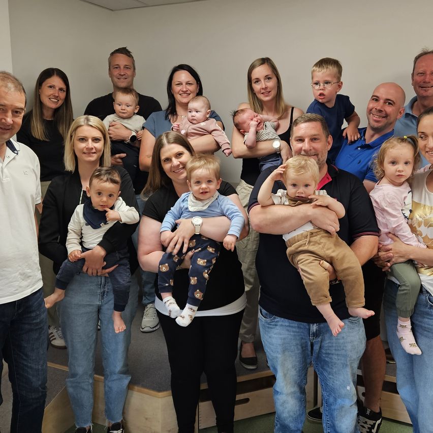 Eine Gruppe von Menschen posiert für ein Foto mit ihren Babys, einschließlich Erwachsener und Kleinkinder, alle lächelnd und ihre Babys haltend. Sie stehen in einem Raum mit weißen Wänden und einem Holzboden.