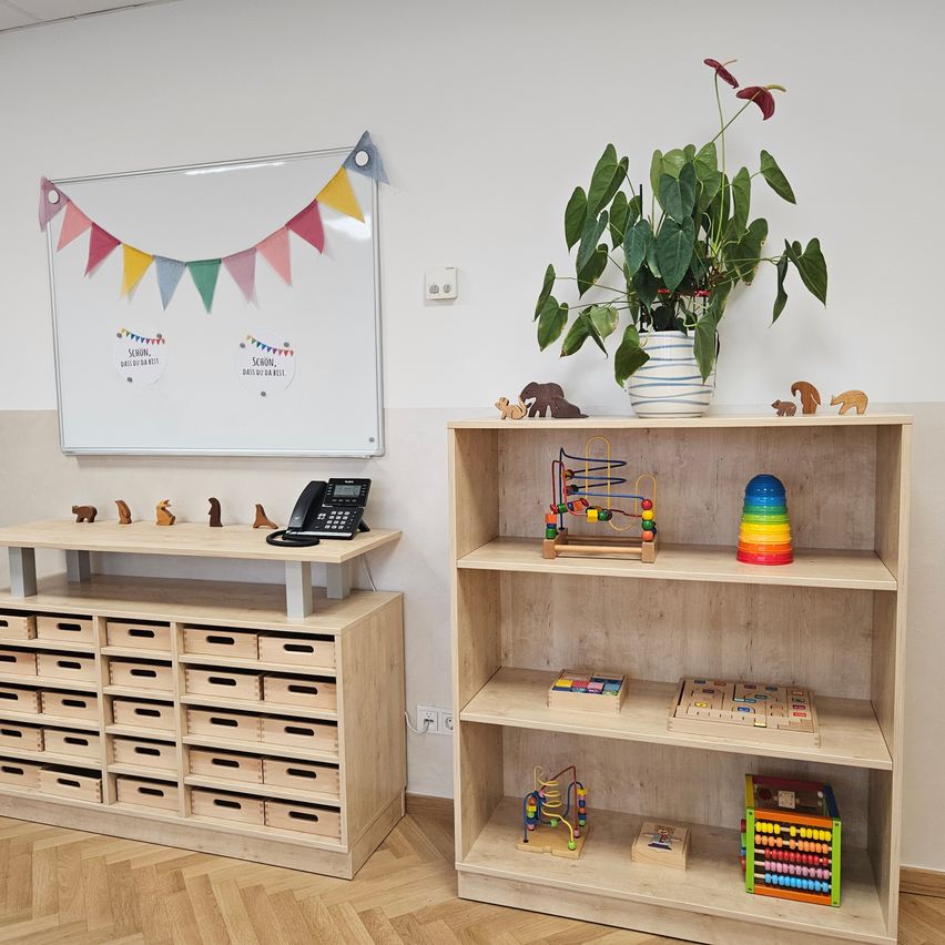 A room with a white wall, a whiteboard with colorful flags, a wooden cabinet with drawers, a telephone, and a shelf with toys and a potted plant.