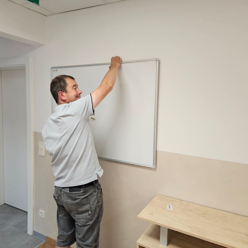 A man in a white shirt and gray shorts is standing in a room and writing on a whiteboard mounted on the wall.