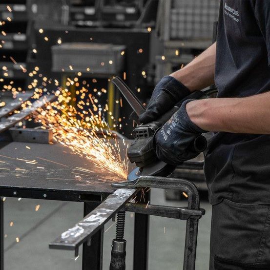 Eine Person verwendet einen Schleifer, um ein Metallblech zu schneiden, wodurch Funken fliegen. Die Person trägt ein schwarzes Shirt und Handschuhe.