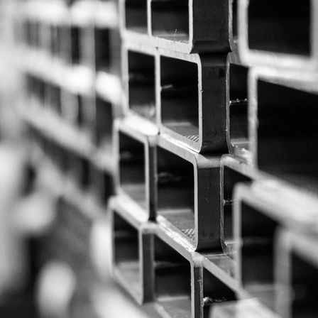 Ein Schwarz-Weiß-Foto eines Stapels von Metallrohren mit quadratischen Öffnungen, möglicherweise in einer Lagerhalle oder Fabrikumgebung.