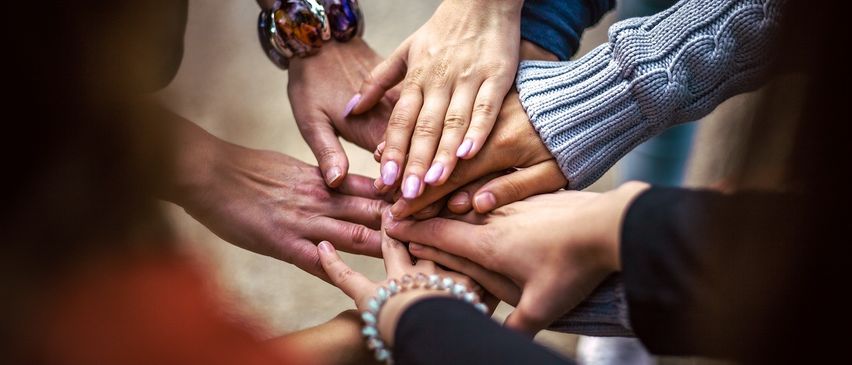 Eine Gruppe von Menschen hat ihre Hände in einem Kreis zusammen, was Einheit und Solidarität zeigt. Sie tragen verschiedene farbige Armbänder.