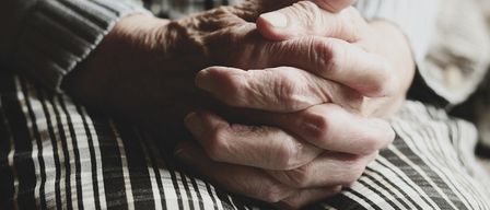 An elderly person sits with their hands clasped together, wearing a long-sleeve shirt and a black and white striped skirt.
