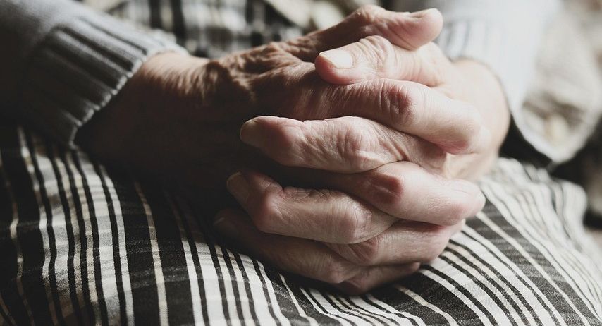 Eine ältere Person sitzt da, die Hände ineinander verschränkt, auf dem Schoß. Sie trägt ein langärmeliges Hemd und einen gemusterten Rock.