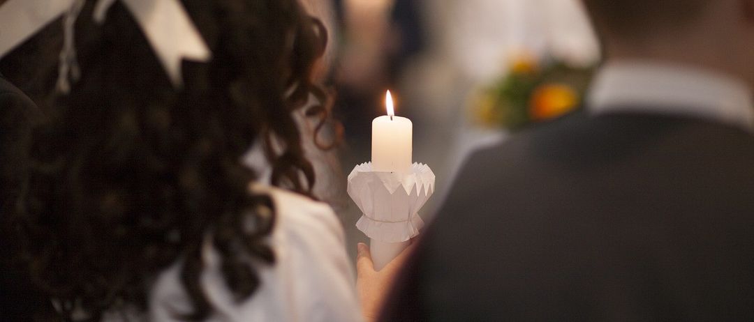 Ein Mädchen in einem weißen Kleid hält eine brennende Kerze bei einer Zeremonie, mit verschwommenen Gästen im Hintergrund.