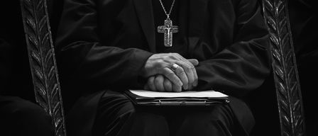 Ein Priester sitzt, trägt einen schwarzen Mantel und hat die Hände gefaltet, während er nach unten schaut. Er trägt eine große Kreuzkette.