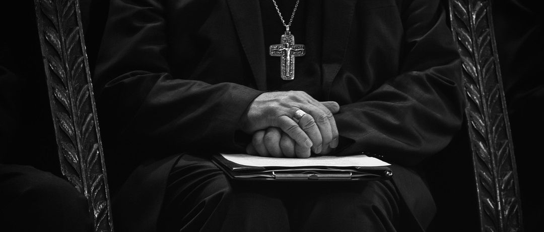 Ein Priester sitzt, trägt einen schwarzen Mantel und hat die Hände gefaltet, während er nach unten schaut. Er trägt eine große Kreuzkette.