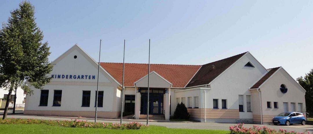 Ein Kindergartengebäude mit rotem Dach und blauem Schild. Es hat einige Fenster und eine Tür. Die Vorderseite hat Gras und Blumen.