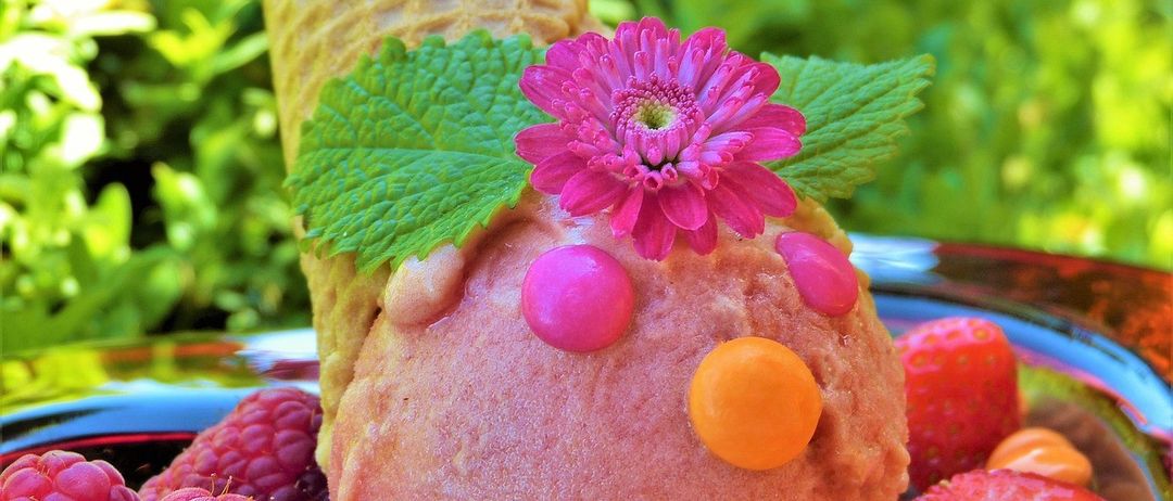 Ein rosafarbener Eisbecher in Gesichtsform ist mit einer rosa Blume, grünen Blättern und bunten Süßigkeiten verziert. Der Kegel steht auf einem Teller mit Himbeeren und Erdbeeren.