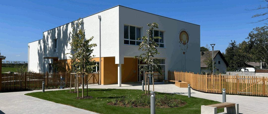 Ein weißes Gebäude mit gelben Akzenten verfügt über einen Garten und einen Holzzaun. Davor stehen zwei Bäume und eine Bank. Das Gebäude hat ein Logo an der Seite.