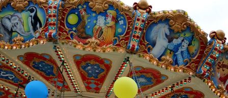 Ein Karussell mit bunten Luftballons und kunstvollen Dekorationen. Ein Clown und ein Pferd sind auf dem Karussell abgebildet. Es hat blaue, gelbe und rote Designs.