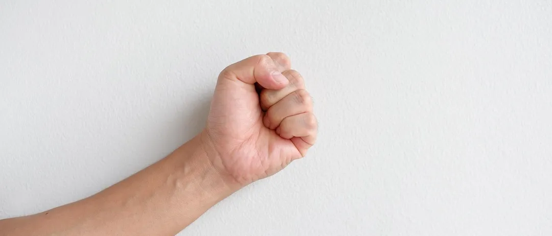 Eine geballte Faust wird gegen eine weiße Wand erhoben und zeigt die Venen am Handgelenk.
