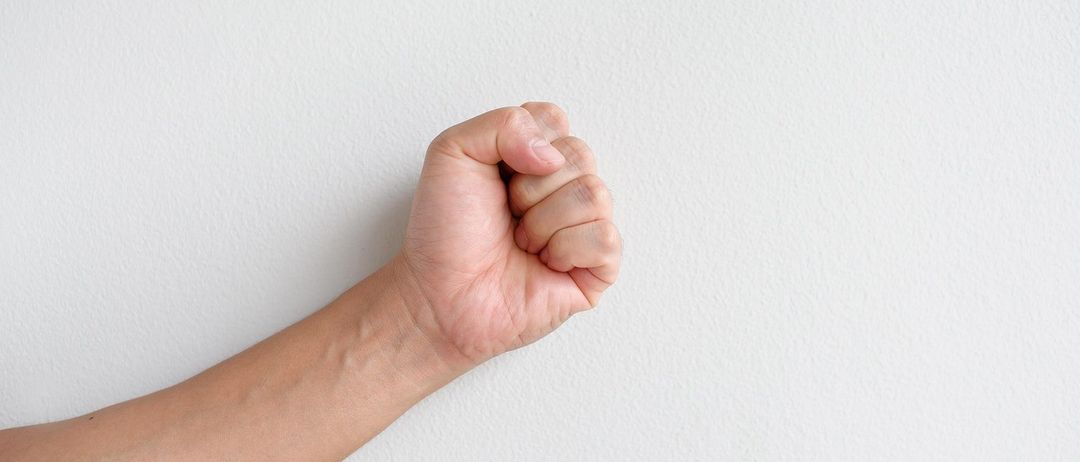 Eine geballte Faust wird gegen eine weiße Wand erhoben und zeigt die Venen am Handgelenk.