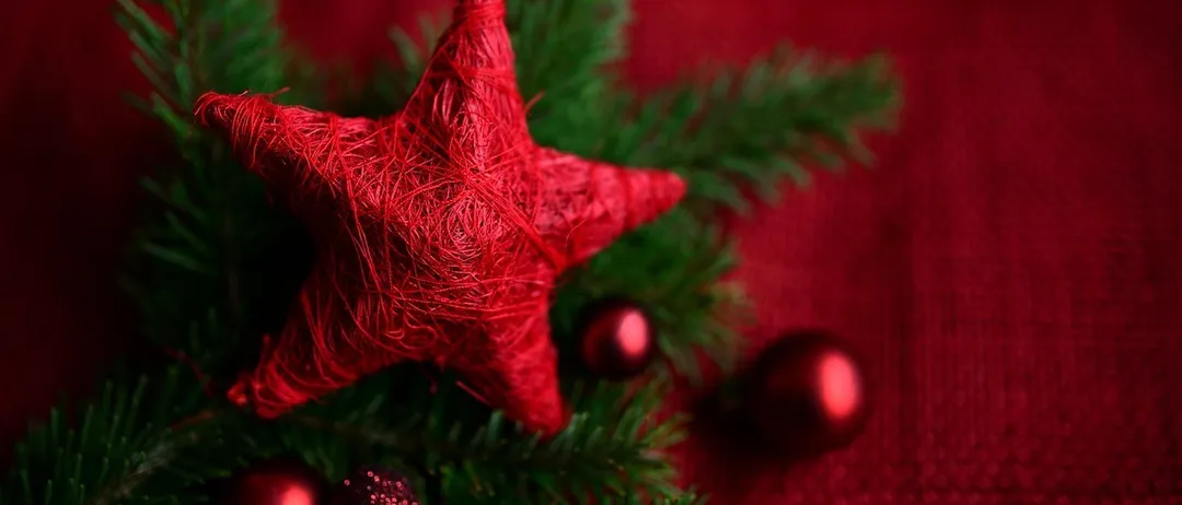 Ein rotes Sternenornament liegt auf rotem Hintergrund mit roten Weihnachtskugeln und grünen Blättern.