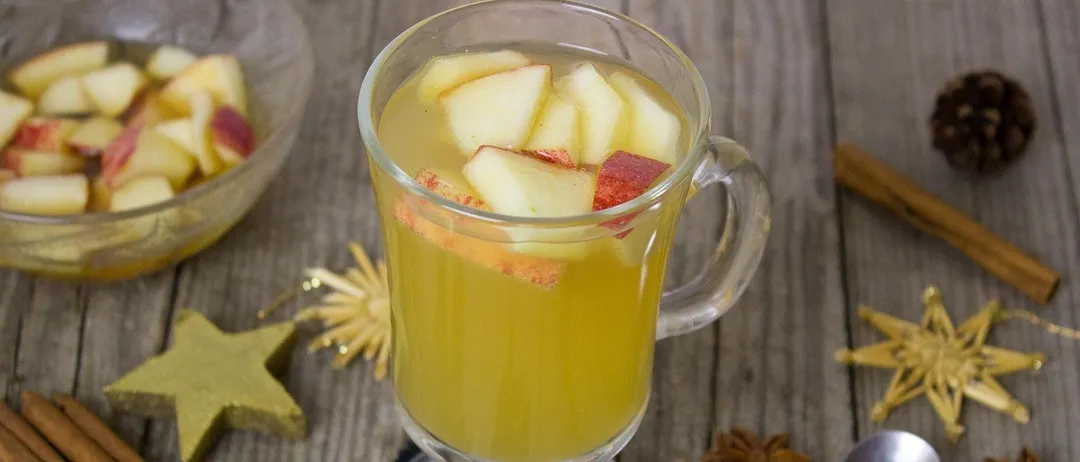 Ein Glas mit Apfelwein, verziert mit Apfelscheiben, auf einem Holztisch mit Tannenzapfen und Sterndekorationen.