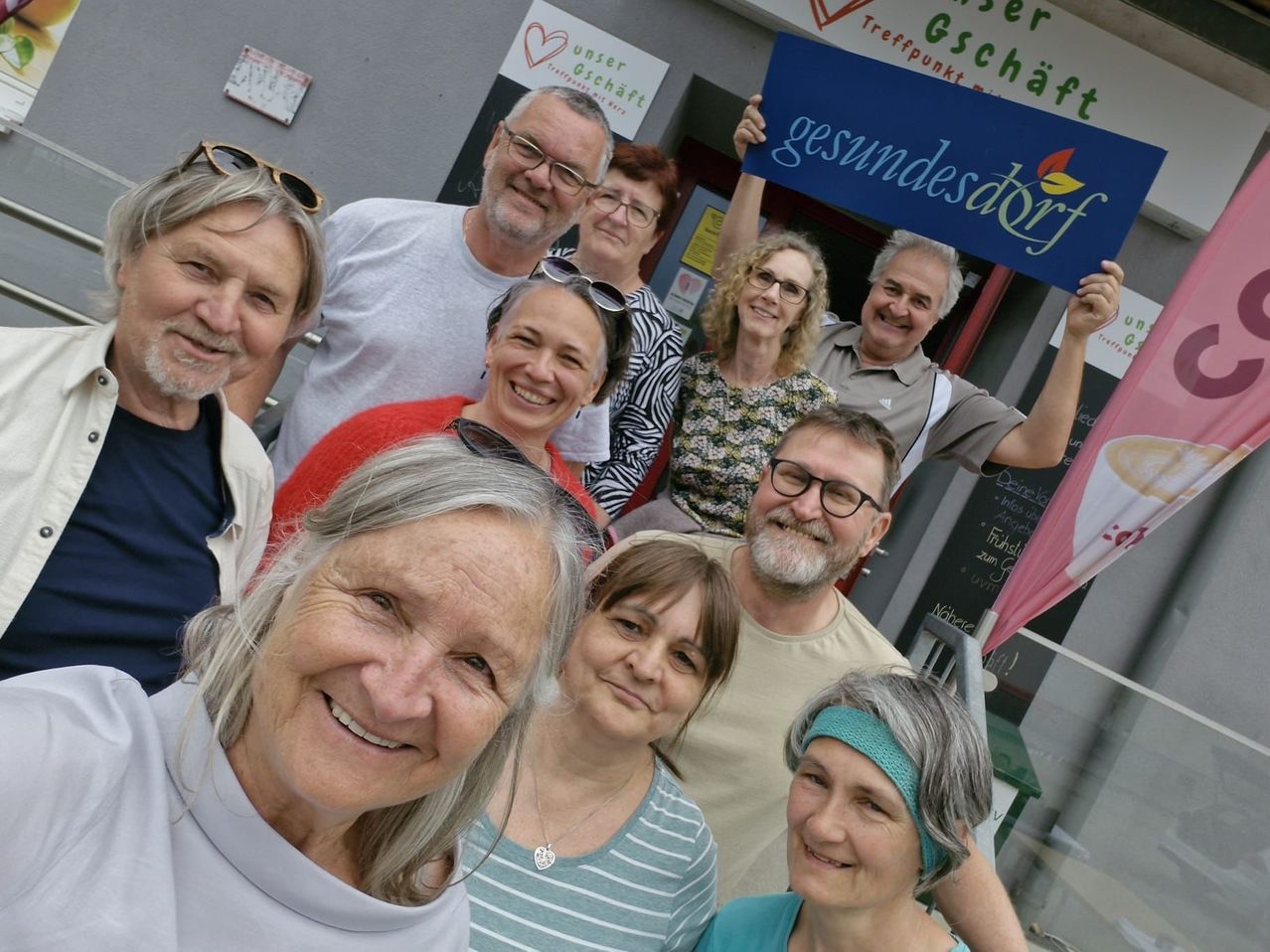 Eine Gruppe von Menschen steht vor einem Gebäude mit einem Schild, auf dem gesundesdorf steht. Sie lächeln und scheinen für ein Foto zu posieren. Einige tragen Brillen und manche haben die Hände erhoben. Es gibt eine Flagge und eine Tafel mit Schrift.