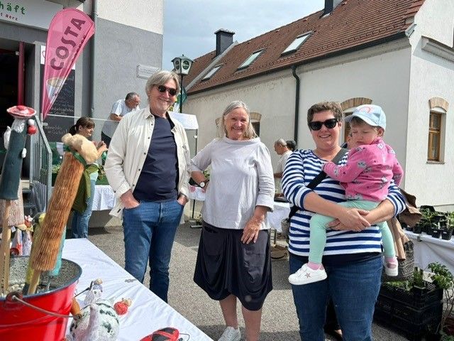 Drei Erwachsene und ein junges Mädchen stehen neben einem Tisch mit verschiedenen Gegenständen. Der Mann trägt eine Sonnenbrille und eine Uhr, und die Frau hält das Mädchen auf der Schulter. Dahinter steht ein Haus mit einem roten Dach und einer Fahne auf der linken Seite.