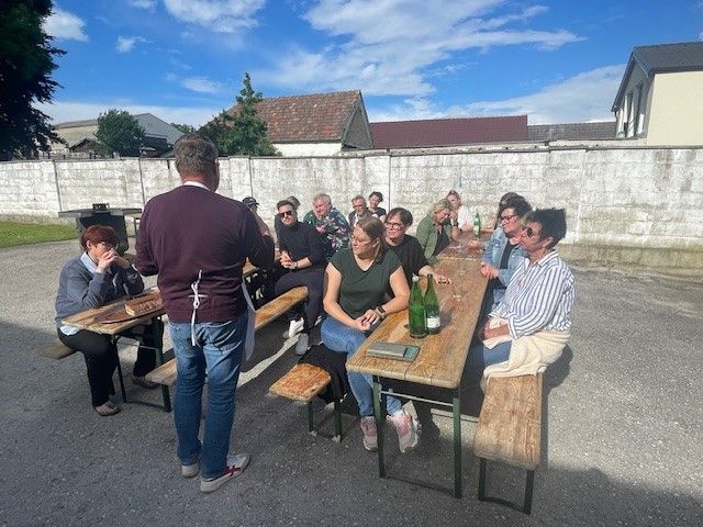 Eine Gruppe von Menschen sitzt an einem Picknicktisch im Freien, einer steht. Flaschen, Gläser und andere Gegenstände sind auf dem Tisch. Eine Frau trägt eine Brille und eine Uhr. Hinter ihnen ist eine Wand und einige Bäume.