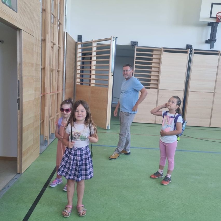 Drei junge Mädchen und ein erwachsener Mann stehen in einer Turnhalle. Die Mädchen tragen Sonnenbrillen und tragen Rucksäcke. Der Mann trägt eine Brille und Turnschuhe. Holztüren sind hinter ihnen offen.